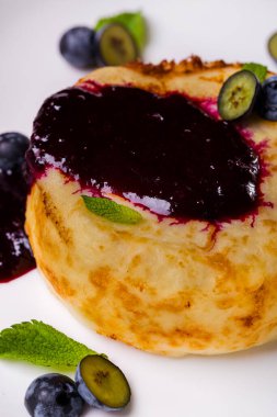 Frenk üzümlü peynirli kek. Yaban mersini ve nane aromalı beyaz bir tabakta yatıyor. Tabak beyaz bir masa örtüsünün üzerinde, iki bardak bir sapın üzerinde, biri meyve suyu ile, diğeri de suyla. Yakınlarda bir çatal ve bıçak var..