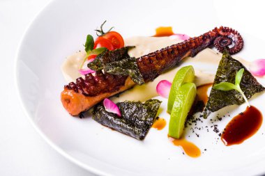 Mantar soslu kızarmış ahtapot, limon dilimleri, unagi sosu, vişneli domates ve mikroyeşil, beyaz tabakta. Çatallı beyaz masa örtüsü, ikinci bir tabak ve bir bardak gül şarabı..