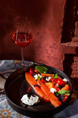 Salad with salmon fillet, tomatoes, basil, feta cheese, caviar and sauce in a plate on the table with a glass of wine against a brown wall