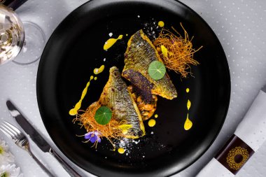 Kızarmış dorado dilimi, tatlı patatesler, dantel cipsler, sarı sos, yuvarlak siyah bir tabakta garnitür, bir bardak şarap ve çatal bıçakla. Yatay yönelim