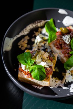 Sandwiches with cheese, meat, basil, almonds, kumquat, sauce and sesame seeds in a black plate against a dark green window background