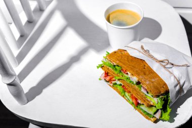 Sandwich in white paper with ham, lettuce, tomatoes, cucumbers and sauce with cappuccino with a paper cup standing on a white chair against the background of a white wooden wall