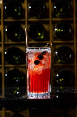 Cold, summer drink lemonade with raspberry syrup. Lemonade, raspberry syrup and ice cubes are poured into a tall, glassy, transparent glass. In a glass, a skewer with blueberries. The glass stands on a black stone round table. Behind the table is a w