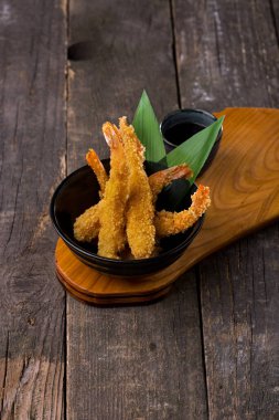 Tempura kaplan karidesi kuyrukları. Karidesler bambu yapraklı siyah bir kasede. Yakınlarda küçük, siyah bir kâse teriyaki sosu var. Kâseler ahşap bir tezgahta duruyor, stantlar tahta bir arka planda duruyor..