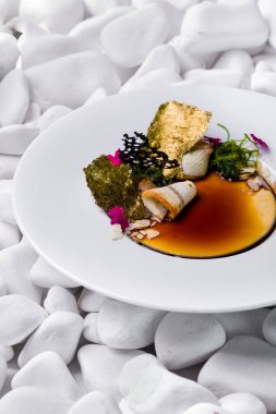 Tütsülenmiş yılan balığı ve balzamik soslu panna cotta, üstünde chuka salatası, badem gevreği, yenilebilir taç yaprakları, yaldızlı Norveç yosunu. Yemek derin beyaz bir tabakta. Tabak beyaz taşların arka planında duruyor..