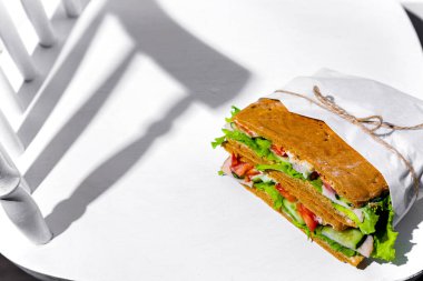 Sandwich in white paper with ham, lettuce, tomatoes, cucumbers and sauce standing on a white chair against the background of a white wooden wall
