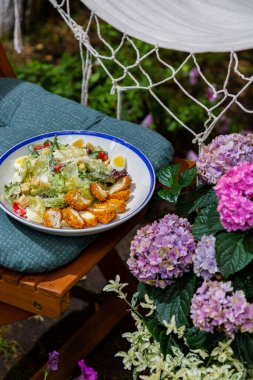 Salad with nuggets, parmesan, sauce, boiled eggs, tomatoes, frisse salad and croutons in a plate on a chair against a background of flowers and herbs