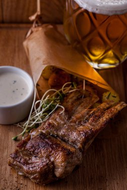 Grilled pork ribs with baked potatoes and microgreen pea sprouts, lie in a paper envelope. Next to it is a white gravy boat with garlic sauce. The food and sauce are on a wooden board, next to a large glass of light beer.