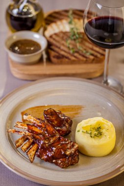 Grilled rack of lamb with barbecue sauce. Next to it is mashed potatoes with butter and dill. The food is on a round ceramic plate with sauce smeared on the plate. The plate is on a beige tablecloth, next to it is a wooden board with grilled toast, t