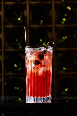 Cold, summer drink lemonade with raspberry syrup. Lemonade, raspberry syrup and ice cubes are poured into a tall, glassy, transparent glass. In a glass, a skewer with blueberries. The glass stands on a black stone round table. Behind the table is a w