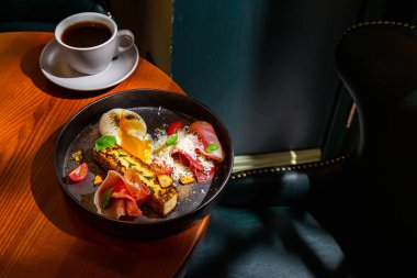 A slice of cheesecake with thyme, prosciutto, parmesan, basil, tomatoes, garlic and a boiled egg in a plate on the table with a cup of coffee