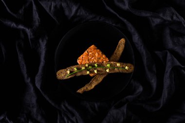 Kare şeklinde hazırlanmış otlu somon tartartarı. Yanında iki çavdar ekmeği, bir yeşil soğan kabuğu ve üzerinde avokado dilimleri var. Yemek siyah yuvarlak bir tabakta. Tabak siyah keten kumaşın üzerinde..