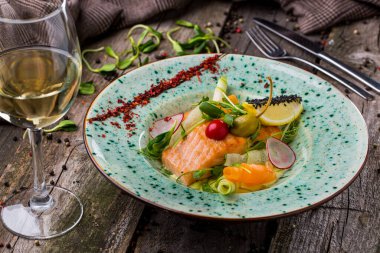 Kavrulmuş somon balığı, fasulye salatası, kiraz domatesleri, salatalık, havuç, turp, limon, siyah susam tohumu, karabiber ve yuvarlak bir tabakta taze bezelye filizi, ahşap bir zemin üzerinde bir kadeh şarap. Yatay yönelim