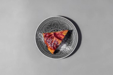 Üzerinde erik dilimleri olan üçgen bir parça erikli kek. Pasta şekerli puding serpiştirilmiş. Pastadan bir parça gri, yuvarlak, seramik bir tabakta kalıp şeklinde duruyor. Tabak gri bir arka planda duruyor..