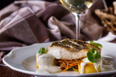 Grilled sea bass steak with white sauce, Korean carrots, mung bean salad, on a white plate with a glass of wine. Horizontal orientation 