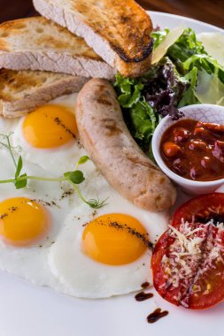 Fried eggs from three eggs with a sprig of microgreen peas, tomatoes with balsamic sauce, lettuce with parmesan slices, three ciabatta toasts and grilled sausage. Nearby is a bowl of beans in tomato sauce. The food is on a white plate. The plate stan