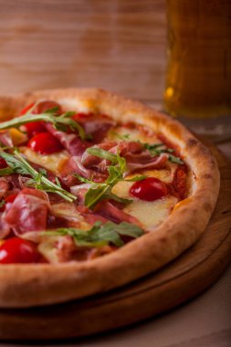 Pizza with cherry tomatoes, prosciutto, arugula and cheese on a wooden board with a glass of beer on the table