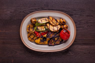 Izgarada ızgara sebze, kırmızı biber, sarı biber, kabak, mantar, soğan, domates, marul yaprağı, zeytinyağı ve otlar. Seramik, oval bir tabakta, ahşap bir arka planda yatıyorlar..