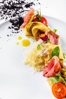 Pancakes with prosciutto, cheese, tomatoes, basil, green pea sprouts, flower petals and white sauce in a plate on a white plate with black sprinkles