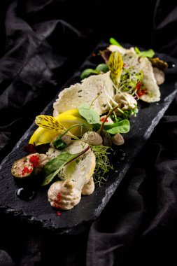 Tavşan ciğeri ezmesi, ekmek cipsi, kapari, marul, balzamik sos, mikroyeşil soğan filizi, limon dilimleri ve yenilebilir yapraklar. Yemek siyah, dikdörtgen, tahta tabakta. Tabak siyah keten bir kumaşın üzerinde..