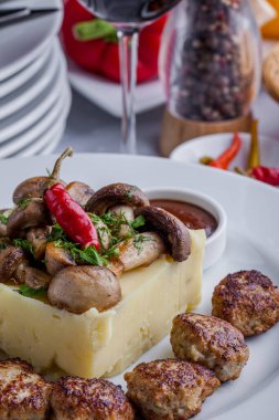 Nine small rabbit cutlets with mashed potatoes and champignons on top, chopped parsley and chilli. Next to the mashed potatoes there is a saucepan with ketchup. The food is on a white plate. The plate is on the table with a white tablecloth, seven em