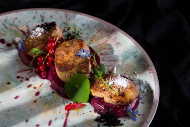 Vişne soslu ızgara kaz ciğeri, buğday ve soğanlı mikro yeşil lahanalar, böğürtlen, ahududu ve yaban mersini, yenilebilir yapraklar. Foie nra yuvarlak bir seramik tabakta yatıyor. Plaka siyah bir arkaplanda duruyor..