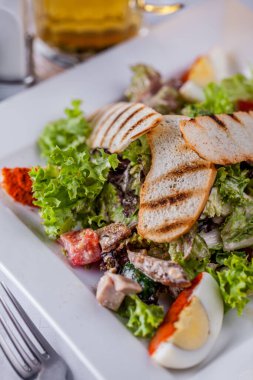 Caesar salad with meat, tomatoes, croutons, lettuce, boiled egg, endive in a plate on a light background with a glass of wine with cutlery