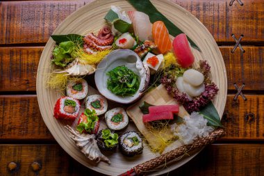 Meat of tuna, shrimp, squid, octopus, salmon, eel, perch with lime, chuka avocado, sesame seeds, carrots, sushi, salmon caviar, cheese, shell, salad and cabbage on banana leaves in a plate on a wooden table and chopsticks