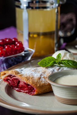 Kirazlı çörek, krema şekeri ve ceviz, ince, kısa hamurlu bir hamur işiyle pişirilmiş. Turta seramik bir tabakta, yanında bir top vanilyalı dondurma ve bir nane yaprağı var. Tabak ahşap bir arka planda duruyor. Yakınlarda kırmızı körili bir kase var.
