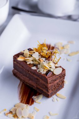 Bir dilim çikolatalı pasta, karamel ve fındık parçalarını beyaz tabakta bir fincan kahveyle birlikte. Dikey yönelim