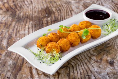 Ten fish croquettes on a white rectangular plate, with onion and pea microgreen. There is a bowl with currant sauce on the plate. The plate stands on a wooden background.