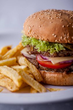 Burger with sesame bun, onion, salad, two meat patties, tomatoes and cheese on a wooden board with fries. Vertical orientation