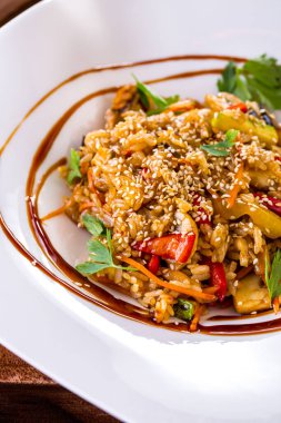 Fried rice with paprika, zucchini, carrots, mushrooms in a wok. Sprinkled with sesame seeds and cilantro leaves with balsamic sauce. The food is on a white, square plate. The plate stands on a wooden background.