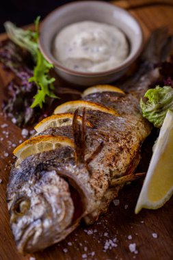 Grilled mackerel stuffed with lemons with lemon slices, white salad dressing and arugula salt on a wooden board. Vertical orientation