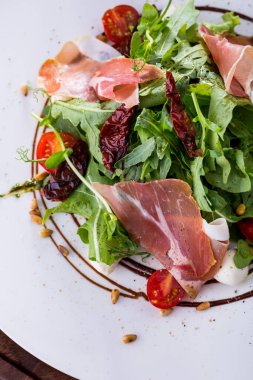 Salad with prosciutto, arugula, tomatoes, lettuce, mozzarella, pea sprouts in a plate with sauce on a wooden background