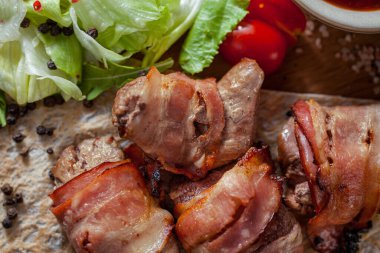 Domuz pastırmasına sarılı ızgara domuz şiş. Şiş kebap pide yatıyor. Yakınlarda dilimlenmiş marul, yarım kiraz domatesleri, tkemali soslu sos kabı ve biraz roka var. Yiyecek tahtanın üzerinde yatıyor. Biberlik ve tuzluk var.