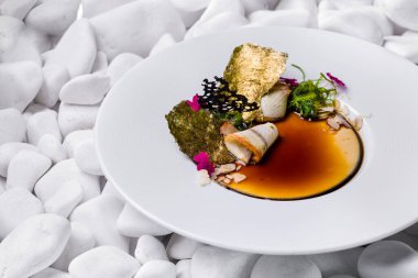 Tütsülenmiş yılan balığı ve balzamik soslu panna cotta, üstünde chuka salatası, badem gevreği, yenilebilir taç yaprakları, yaldızlı Norveç yosunu. Yemek derin beyaz bir tabakta. Tabak beyaz taşların arka planında duruyor..