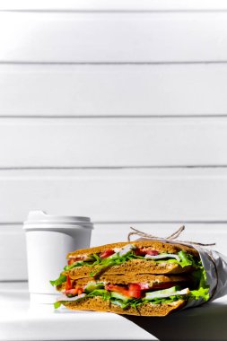 Sandwich in white paper with ham, lettuce, tomatoes, cucumbers and sauce with a paper cup of espresso with a lid standing on a white chair against the background of a white wooden wall