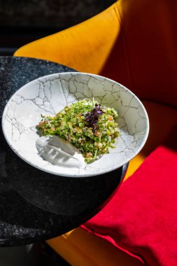 Oriental salad appetizer Tabbouleh with quinoa, finely chopped parsley, cucumber, tomato and microgreens. Tabbouleh lies in a light round ceramic plate with high sides. The plate sits on a black table covered in sparkles. Nearby is a yellow armchair 