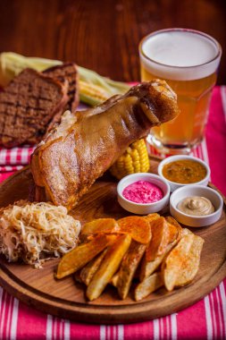 Bir bardak birayla domuz budu, fırında patates, tost, mısır, sos, lahana, ahşap bir masanın üzerinde beyaz çizgili kırmızı bir masa örtüsü ve kırmızı ve beyaz kareli bir peçete.