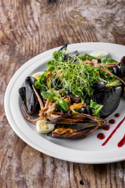 Sarımsak ve bitkilerle tereyağına batırılmış midyeler beyaz, yuvarlak bir tabakta uzanıyorlar. Üstte mikroyeşil roka, ahtapot ve bıldırcın yumurtası salatası var. Vişne sosu tabağa bulaşır. Tabak ahşap bir arka planda duruyor..
