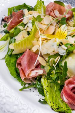 Salad with prosciutto, grapes, lettuce, arugula, grains, almond slices, avocado, flower petals in a plate on a light background 