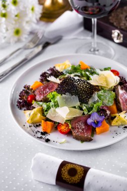 Salad with beef meat, tomatoes, lettuce, cheese, lace chips, endive, carrots, flower petals in a plate on a light background with a glass of wine and cutlery