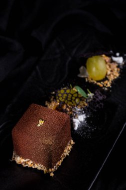 Çikolata parçalı kaz ciğeri ezmesi ve doğranmış ceviz yastığında yenebilir altın. Yakınlarda bir fındık kırıntısında üzüm var. Yemek siyah, dikdörtgen, tahta tabakta. Tabak siyah keten kumaşın üzerinde..