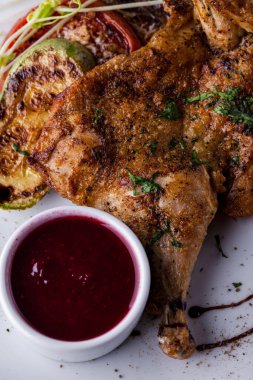 Sarımsak soslu yarım tavuk, doğranmış kişniş, mikroyeşil lahanalar ve balzamik sos. Yanında bir kase ahududu sosu var. Yemek beyaz bir tabakta, ahşap bir masanın üzerinde. Bardakta çorba var, metal bardak tutacağında.