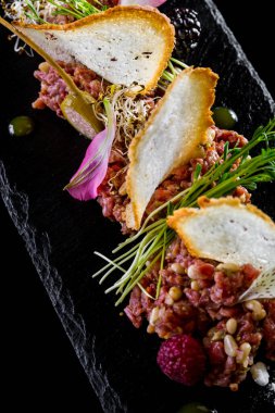 Buğday ekmekli tartar, mikro yeşil mercimek lahanası, kapari, ahududu ve yenilebilir taç yaprakları. Yemekler siyah bir tabakta, tabaklar siyah bir zeminde..