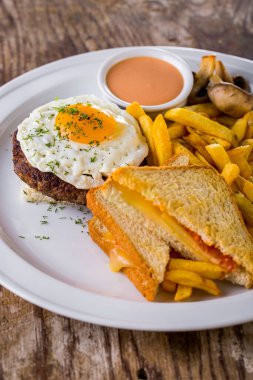 Fried eggs on a pork cutlet with cheese and tomato sandwiches, fried mushrooms, french fries and paprika sauce in a white saucepan. The food is on a white plate.  The plate stands on a wooden background.