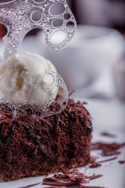 Dessert brownie with chocolate chips, a scoop of ice cream, with transparent icing on a white plate with cream, a cup of coffee and sugar. Vertical orientation
