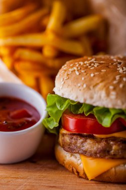 Burger with sesame bun, onion, lettuce, meat cutlet, tomatoes and cheese on a wooden board with sauce and fries. Vertical orientation