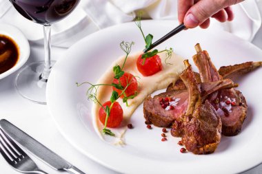 Medium rare veal rack, sliced in portions with mashed potatoes, stewed tomatoes and peppercorns. The food is on a white plate, next to it is barbecue sauce and a glass of red wine. The plate is on a white tablecloth with a fork and knife nearby. The 
